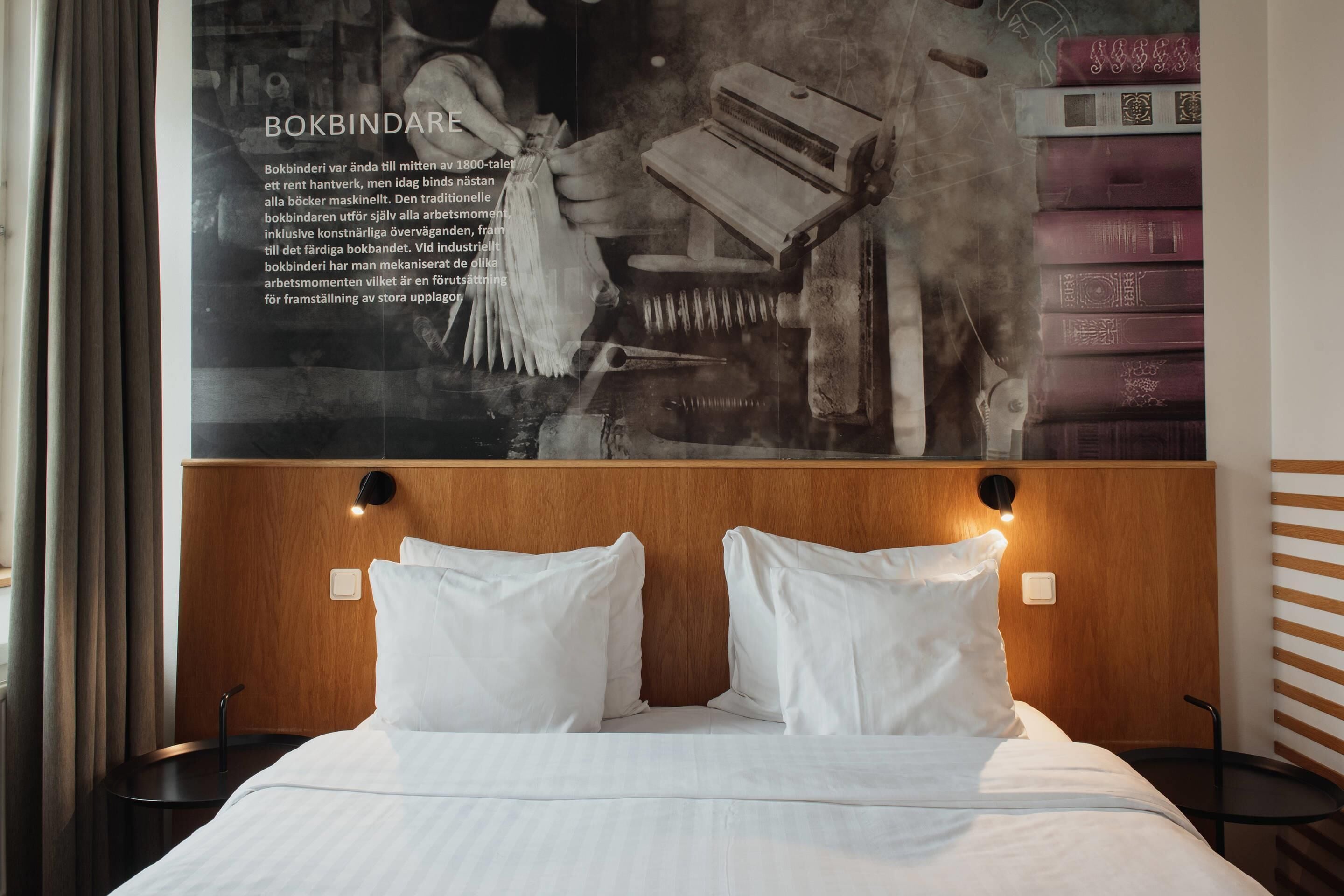 A hotel room with a wooden headboard, two white pillows, a neatly made bed, and a wall-mounted poster with text and images.