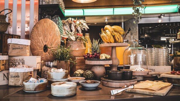 Variedad de alimentos y recipientes a la vista, incluyendo una escultura de pájaro de madera, cestas y diversos tipos de frutas y quesos.
