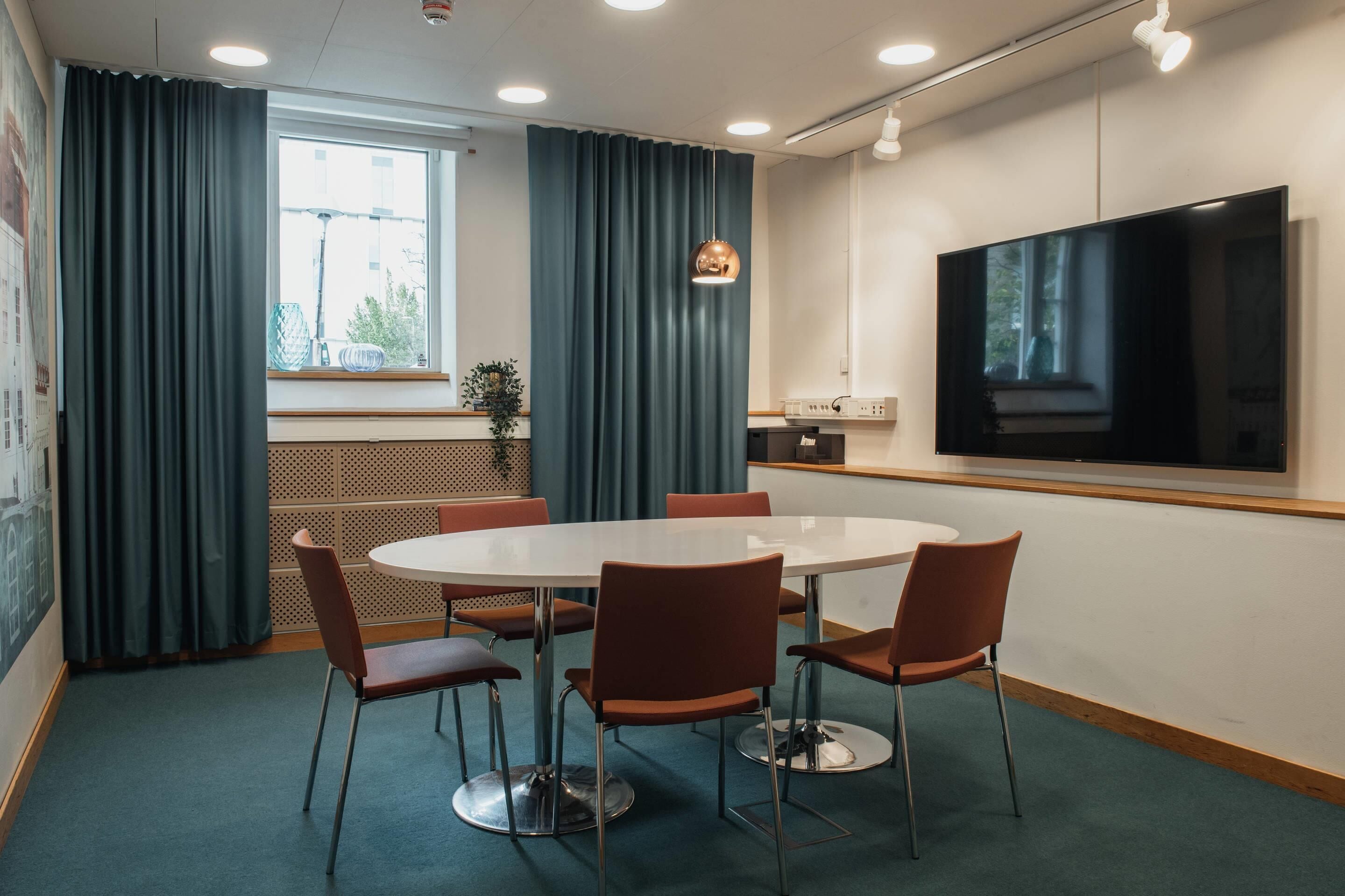 A conference room with a round table, six chairs, a large flat-screen TV, and a window with curtains.
