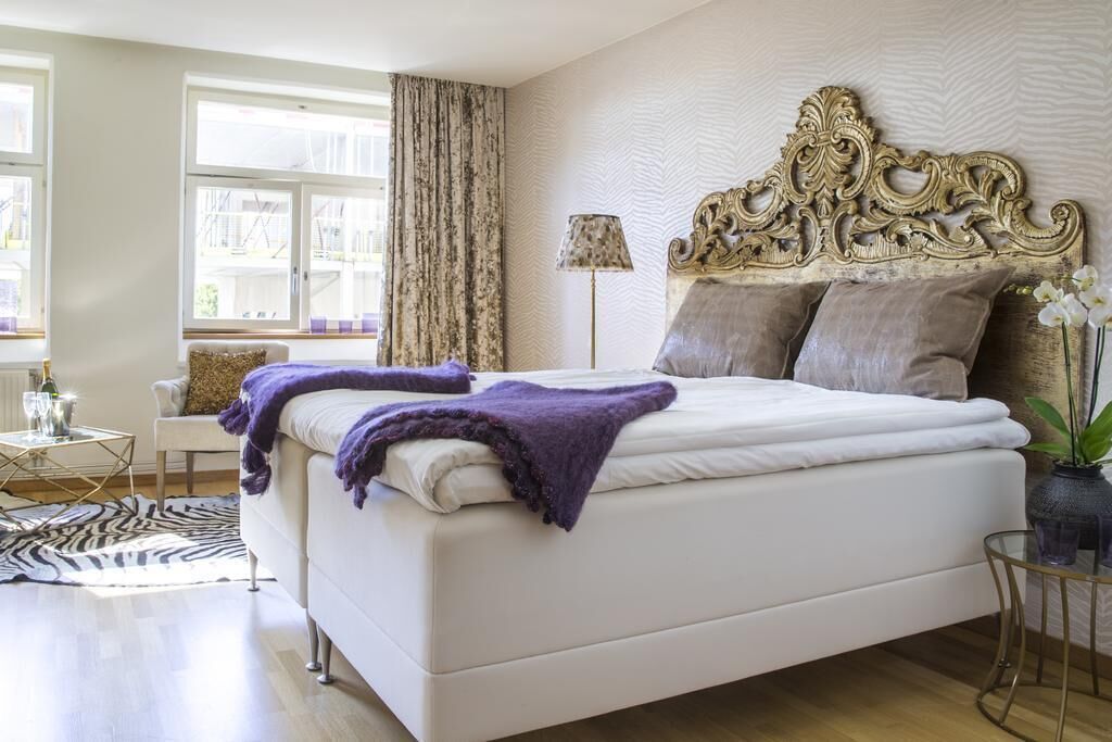 A bedroom with a large bed, a decorative headboard, a small table with a lamp, and a view of the outside through a window.
