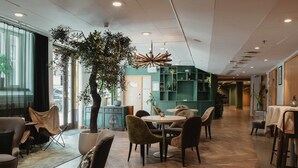 Un interior moderno con piso de madera, mesas redondas y sillas cómodas. Hay plantas en macetas y un árbol grande en el centro.