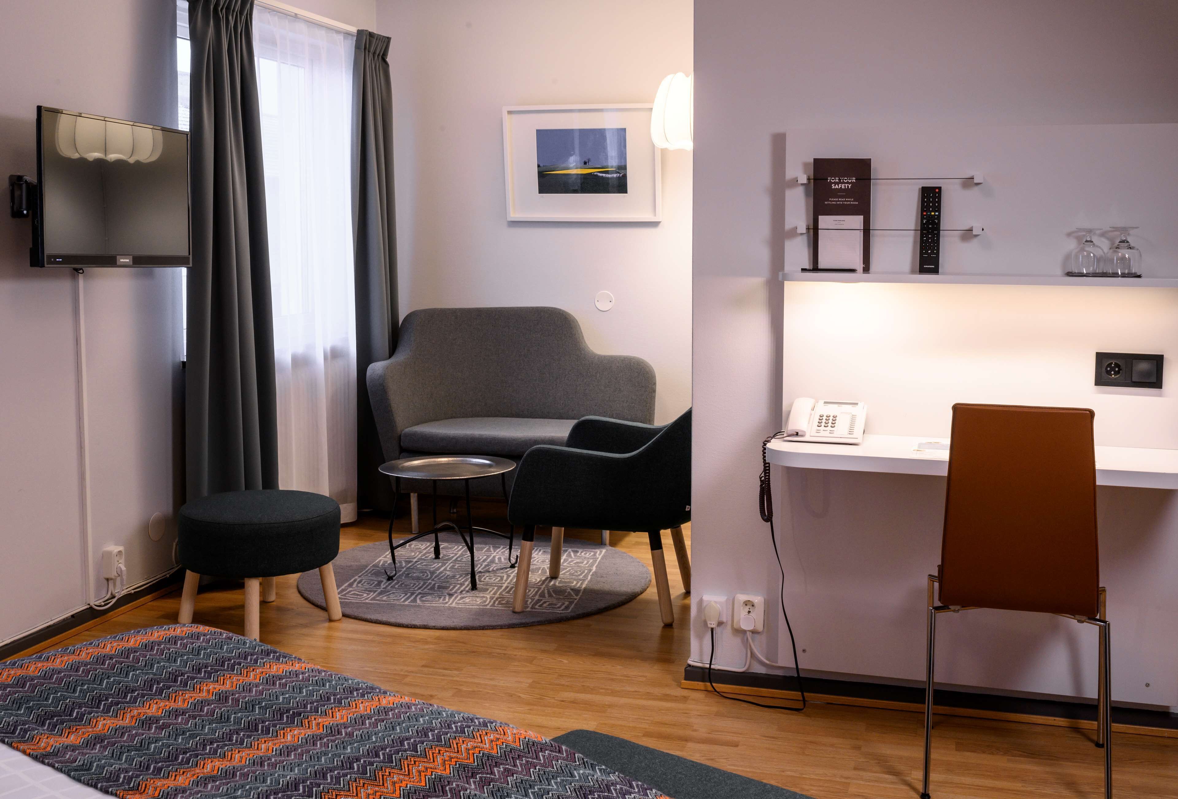 Una camera d'albergo moderna con un divano grigio, un piccolo tavolino rotondo, una sedia marrone, una scrivania con un telefono e un quadro appeso al muro.