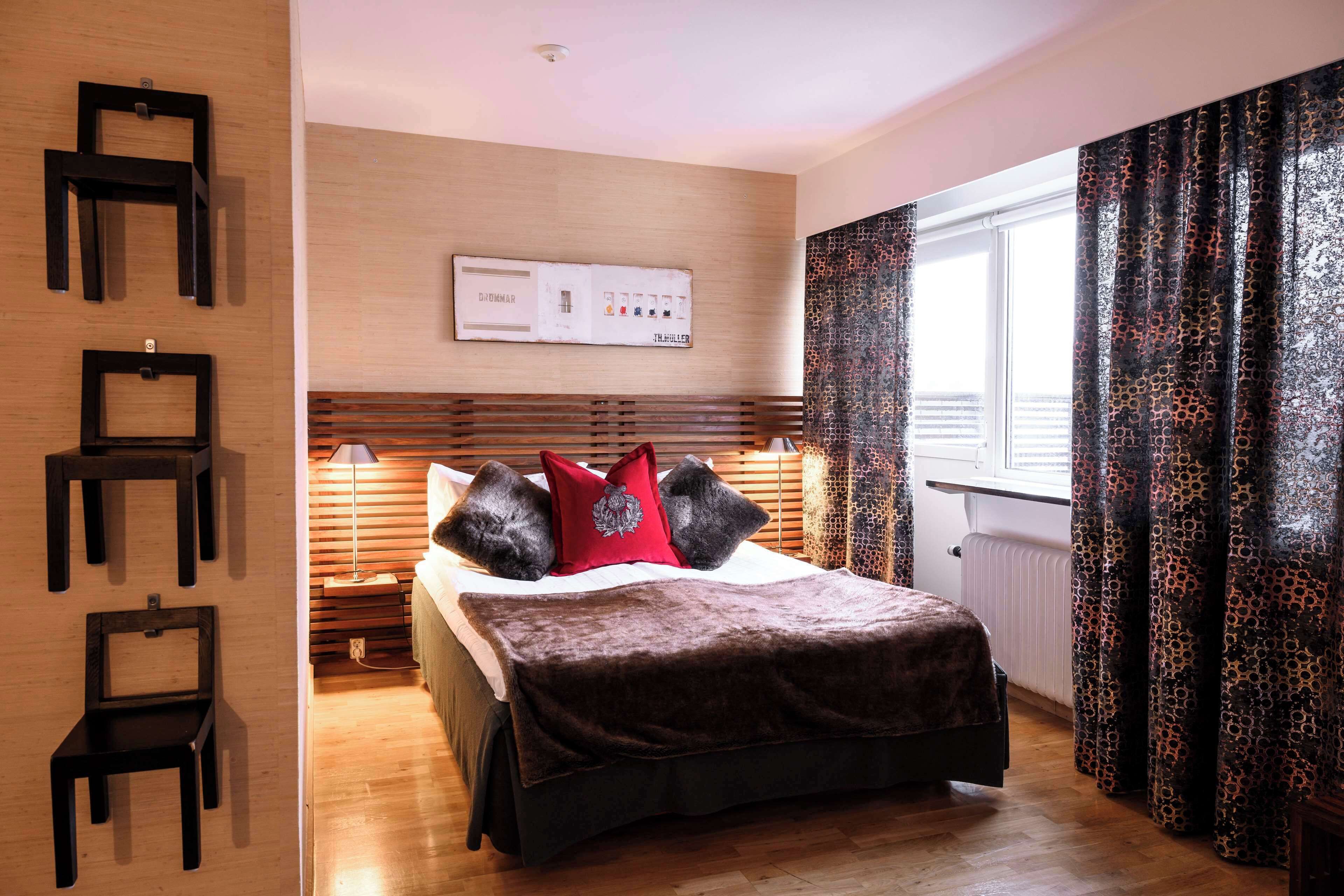 Une chambre à coucher avec une tête de lit en bois, un lit recouvert d’une couverture marron, un oreiller rouge et une étagère fixée au mur, sur laquelle sont posées trois chaises noires.