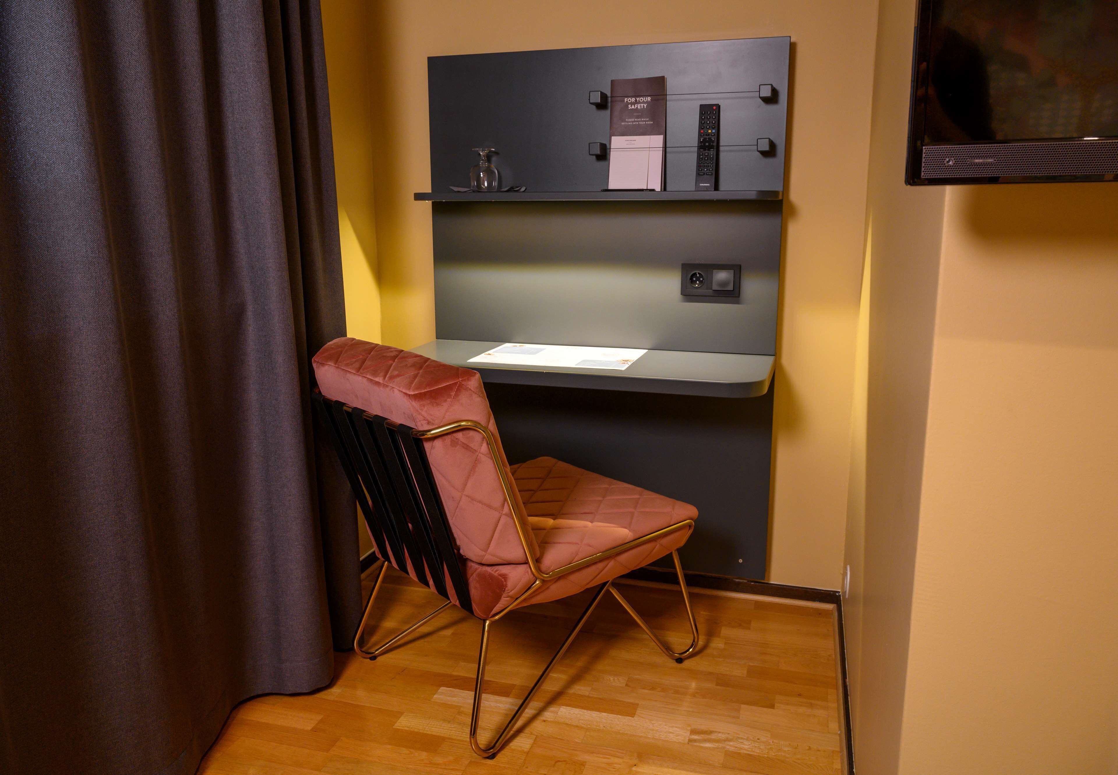 Et værelse med trægulv, en pink polstret stol, et sort skrivebord med en computerskærm, en fjernbetjening og en lille vase.