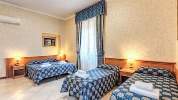 Habitación de hotel con dos camas, cada una con cubrecama floral azul y toallas blancas. Se observa una ventana con cortinas azules, un aire acondicionado de pared y un cuadro enmarcado.