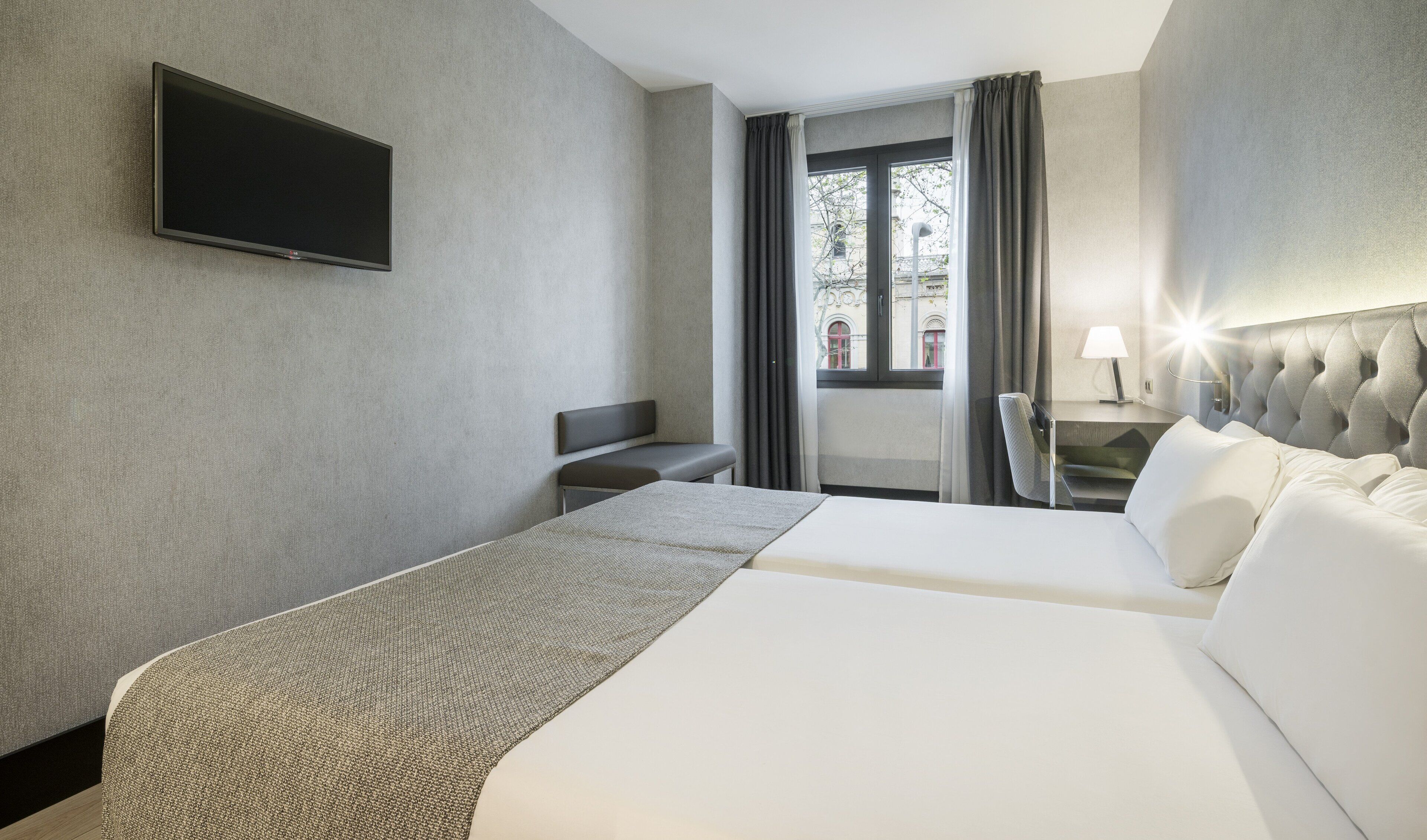 A hotel room with a bed, a desk, a chair, a TV, and a window with curtains.