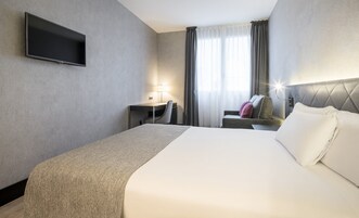 A hotel room with a bed, a desk, a chair, a TV, and a window with curtains.