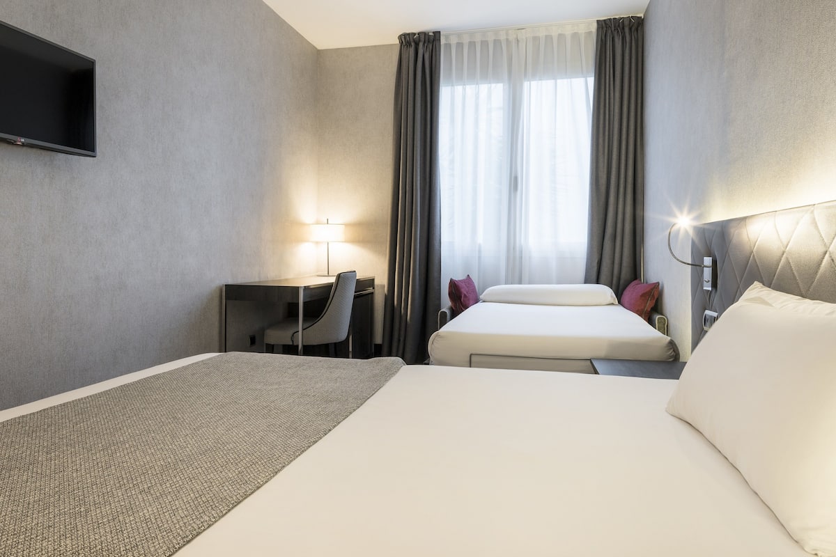 Et hotelværelse med seng, skrivebord, stol, fjernsyn og et vindue med gardiner.
