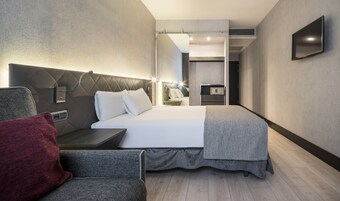 A modern hotel room with a large bed, a bedside table, a chair, and a flat-screen TV mounted on the wall.