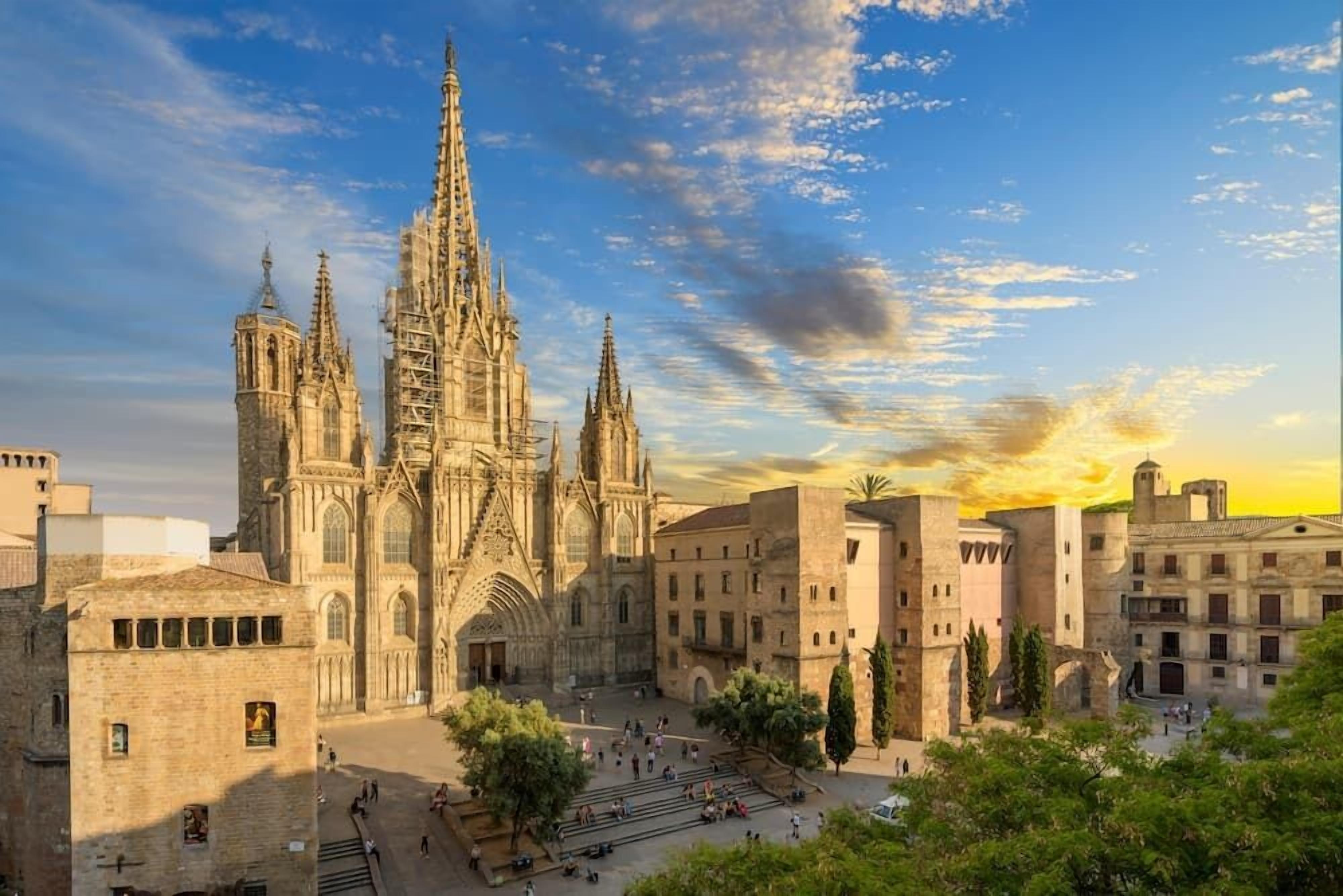 A historic cathedral with a large central spire and multiple towers, surrounded by a courtyard with trees and a few people.