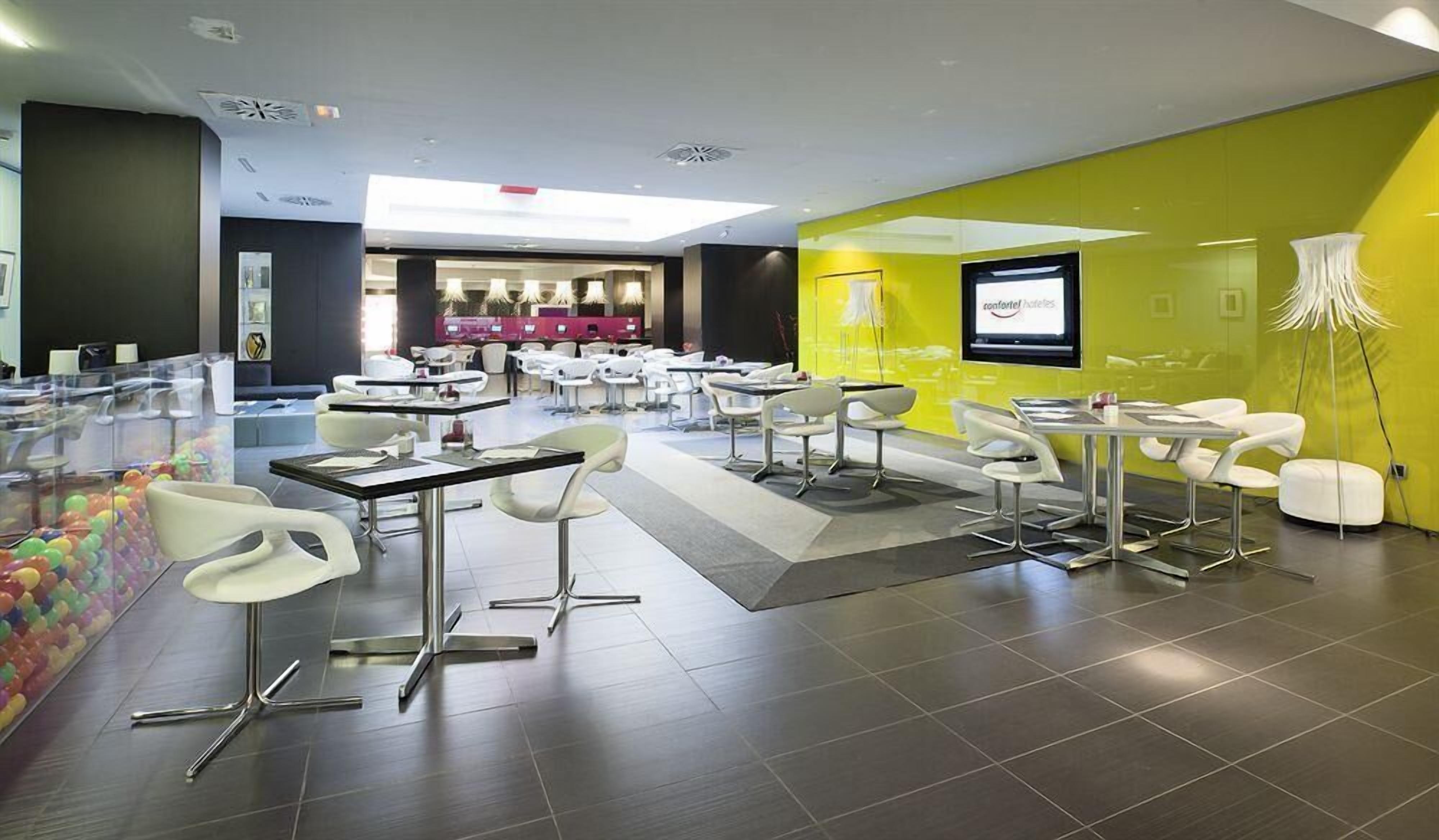 A modern hotel lobby with a bright yellow accent wall, a digital screen displaying the hotel name, and a seating area with white chairs and tables.