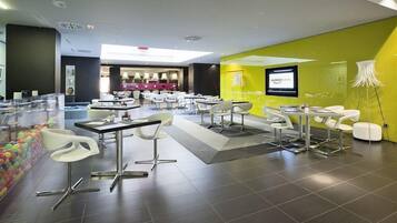 A modern hotel lobby with a bright yellow accent wall, a digital screen displaying the hotel name, and a seating area with white chairs and tables.