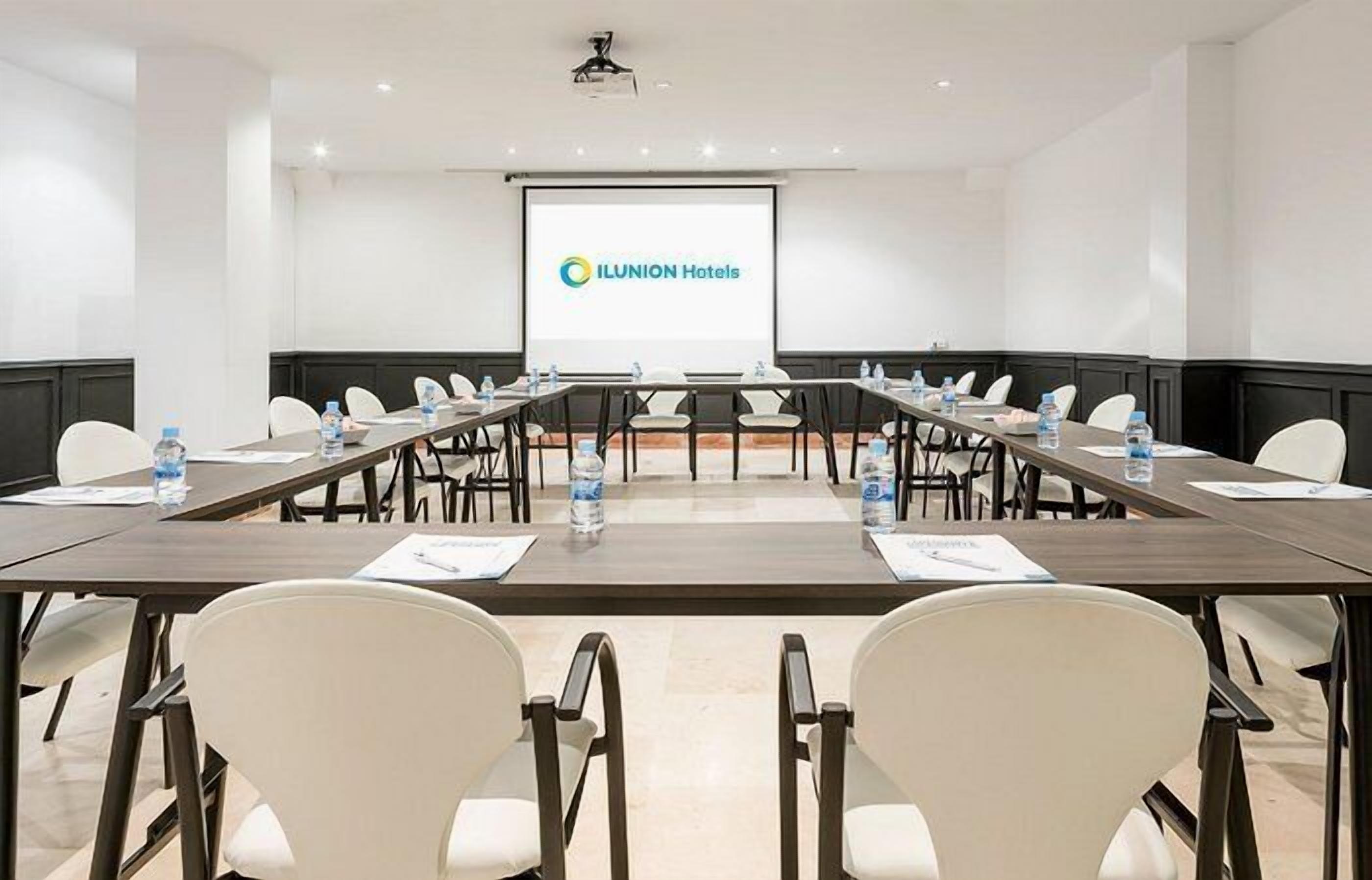 Ein Konferenzraum mit einem langen Tisch, weißen Stühlen und einer Projektionsleinwand mit dem ILUNION Hotels Logo.