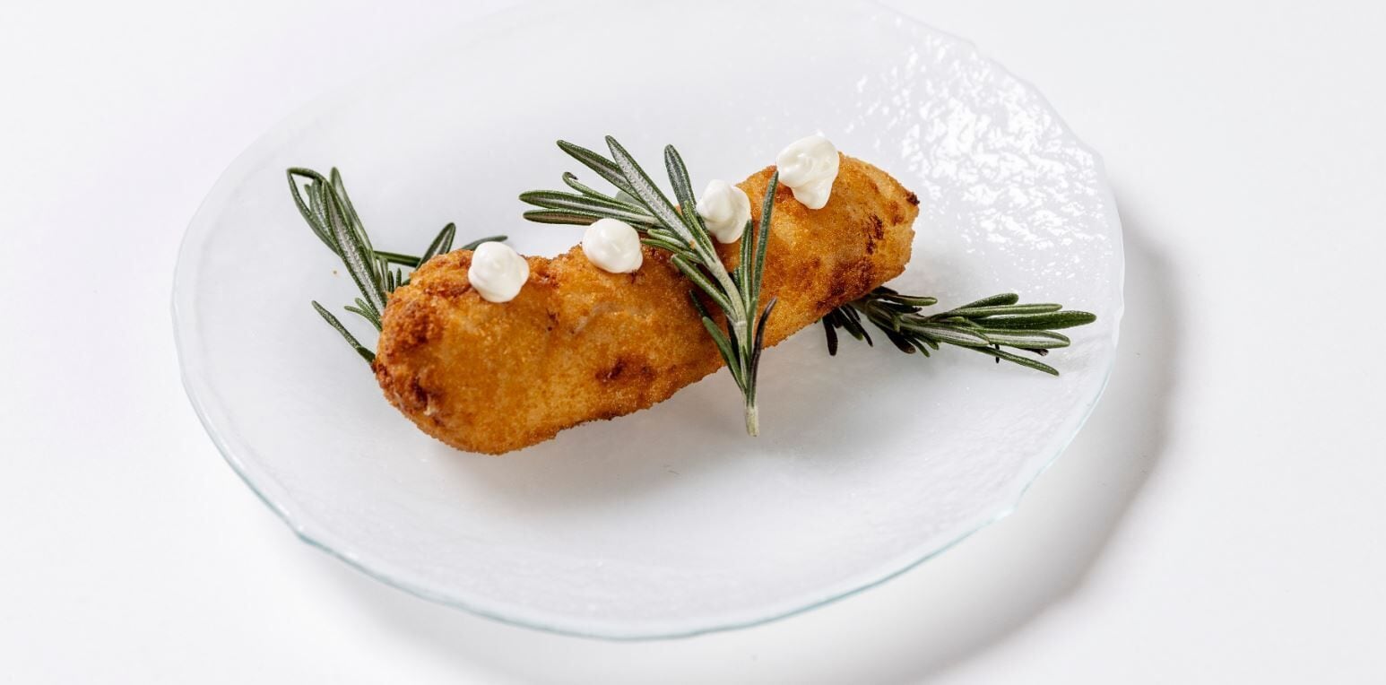 Dos preparaciones rebozadas y fritas sobre un plato blanco, con romero y cucharadas de crema.