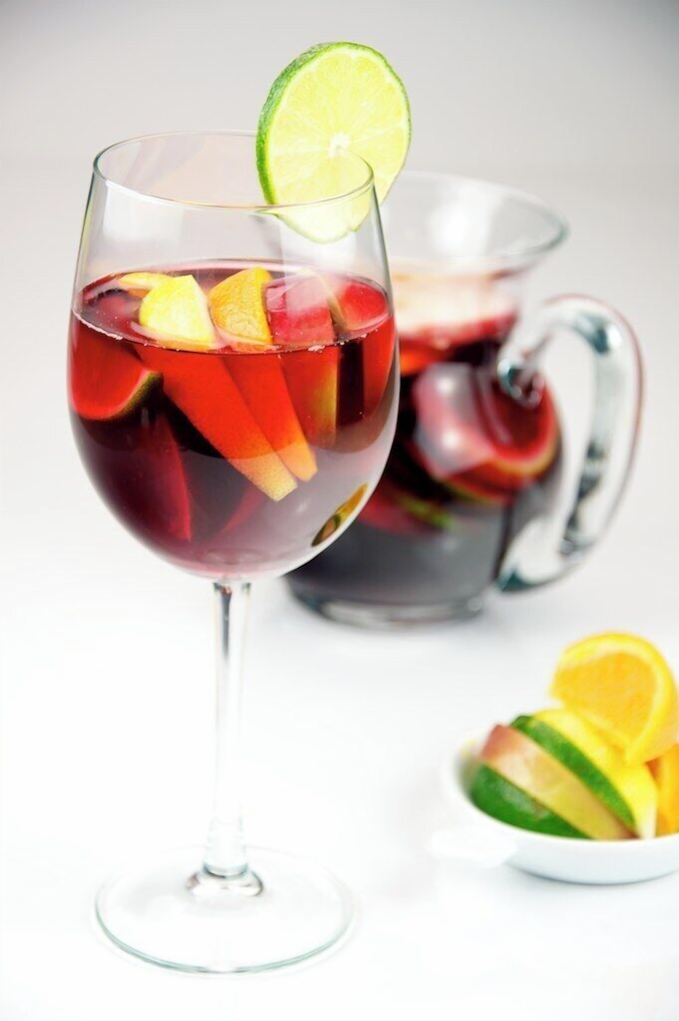 A glass of red drink with fruit slices and a lime wedge, a pitcher of the same drink, and a bowl of fruit in the background.