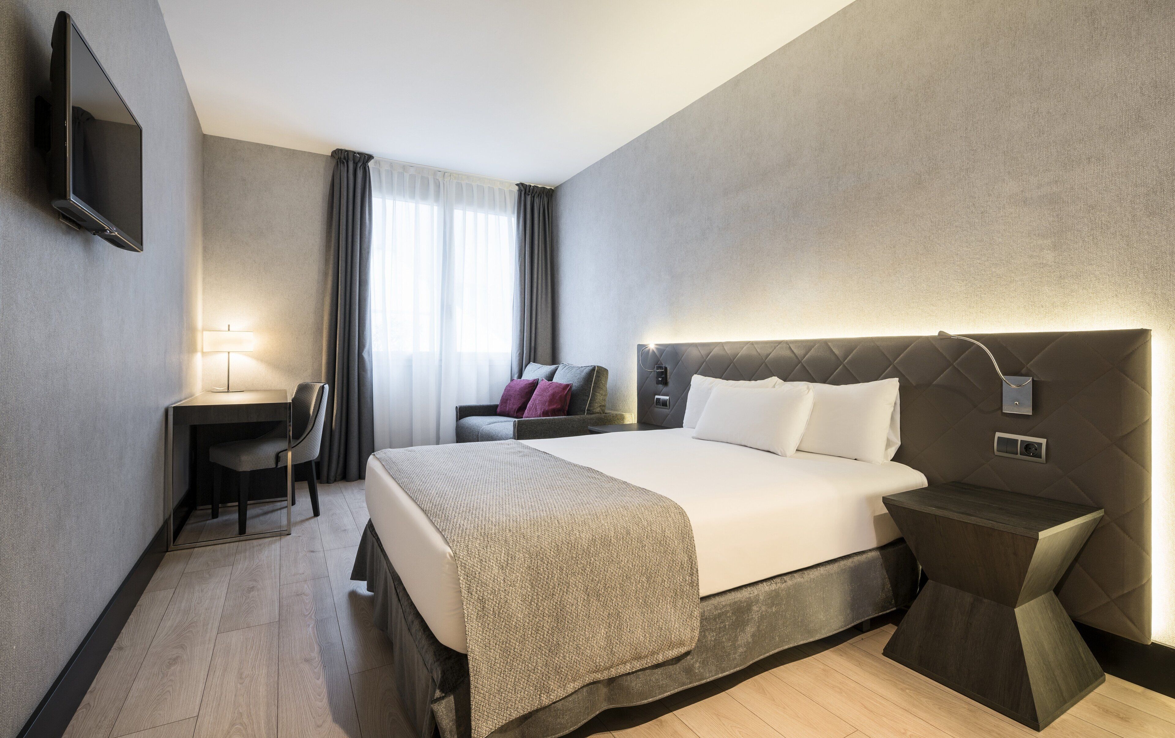 Een hotelkamer met een bed, nachtkastje, lamp, bureau, stoel en een aan de muur bevestigde televisie.