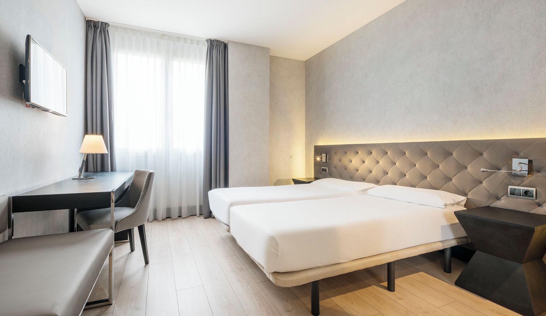 Habitación de hotel con cama, escritorio, silla, lámpara y ventana con cortinas.