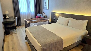 A hotel room with a large bed, a desk with a lamp, a chair, a suitcase, and a nightstand with a lamp.