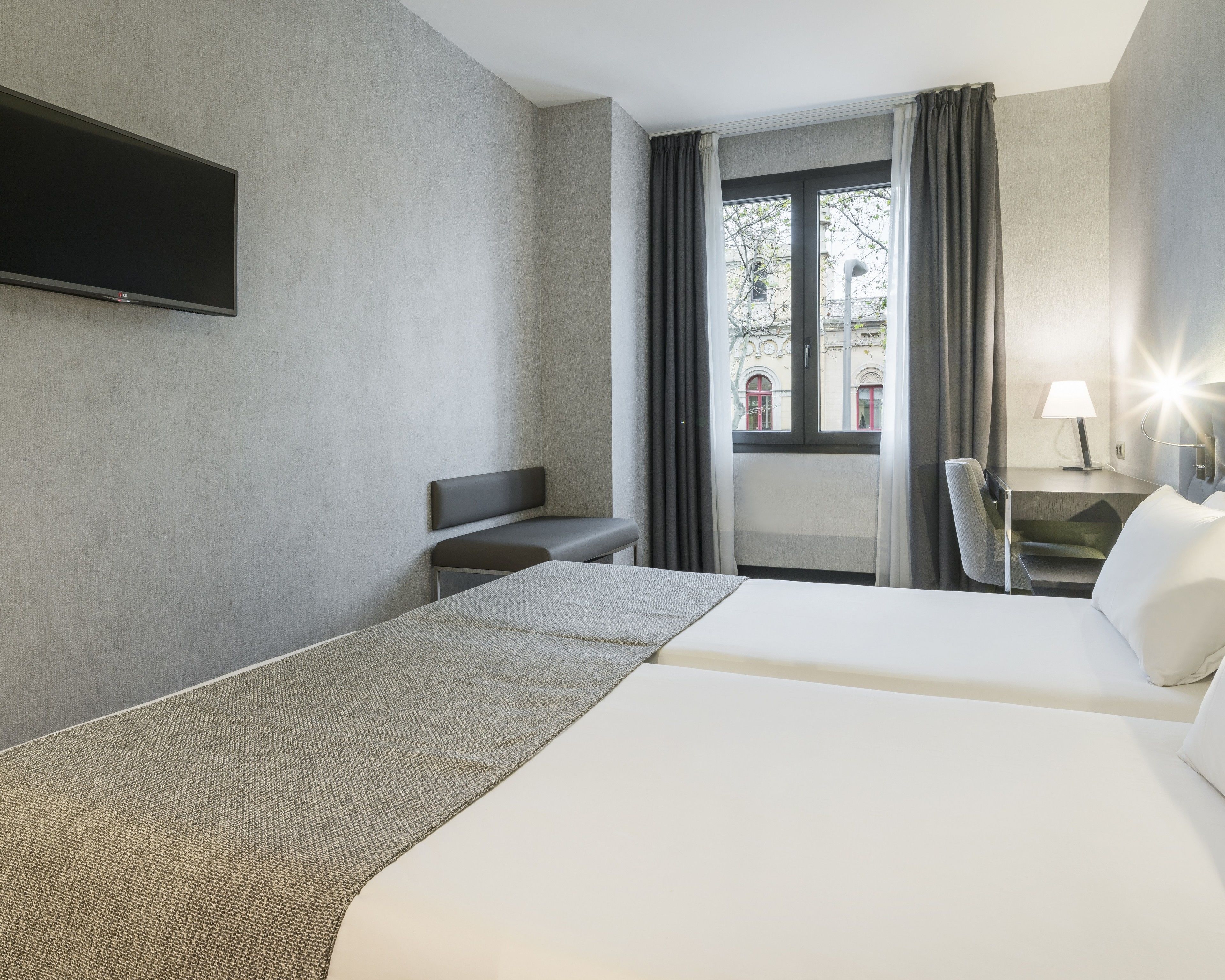 A hotel room with a bed, a bench, a TV mounted on the wall, a window with curtains, and a lamp on a bedside table.