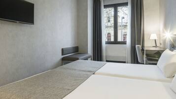Una habitación de hotel con una cama, un banco, un televisor montado en la pared, una ventana con cortinas y una lámpara en una mesita de noche.