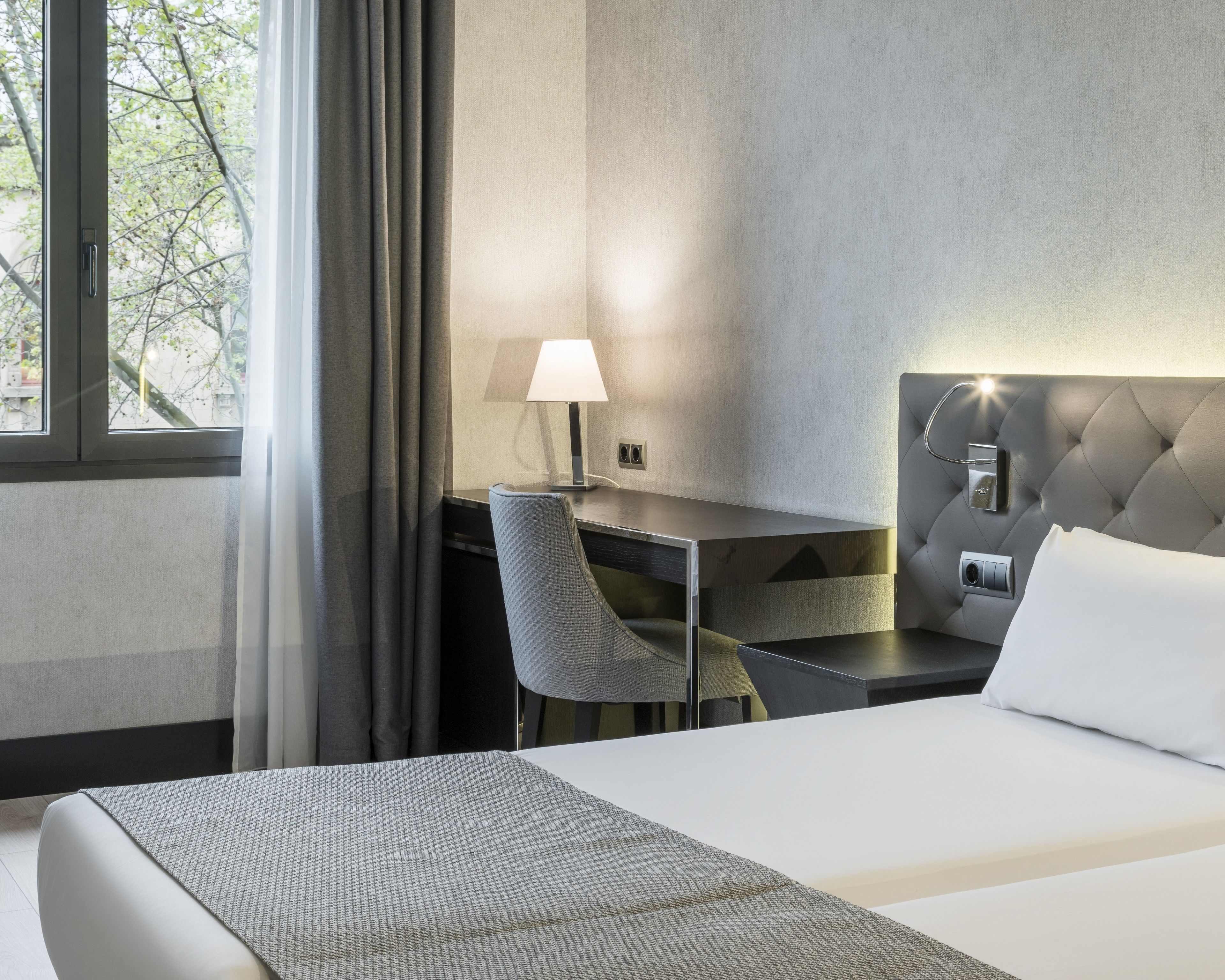 Et hotelværelse med seng, skrivebord, stol, lampe og udsigt til træer gennem vinduet.