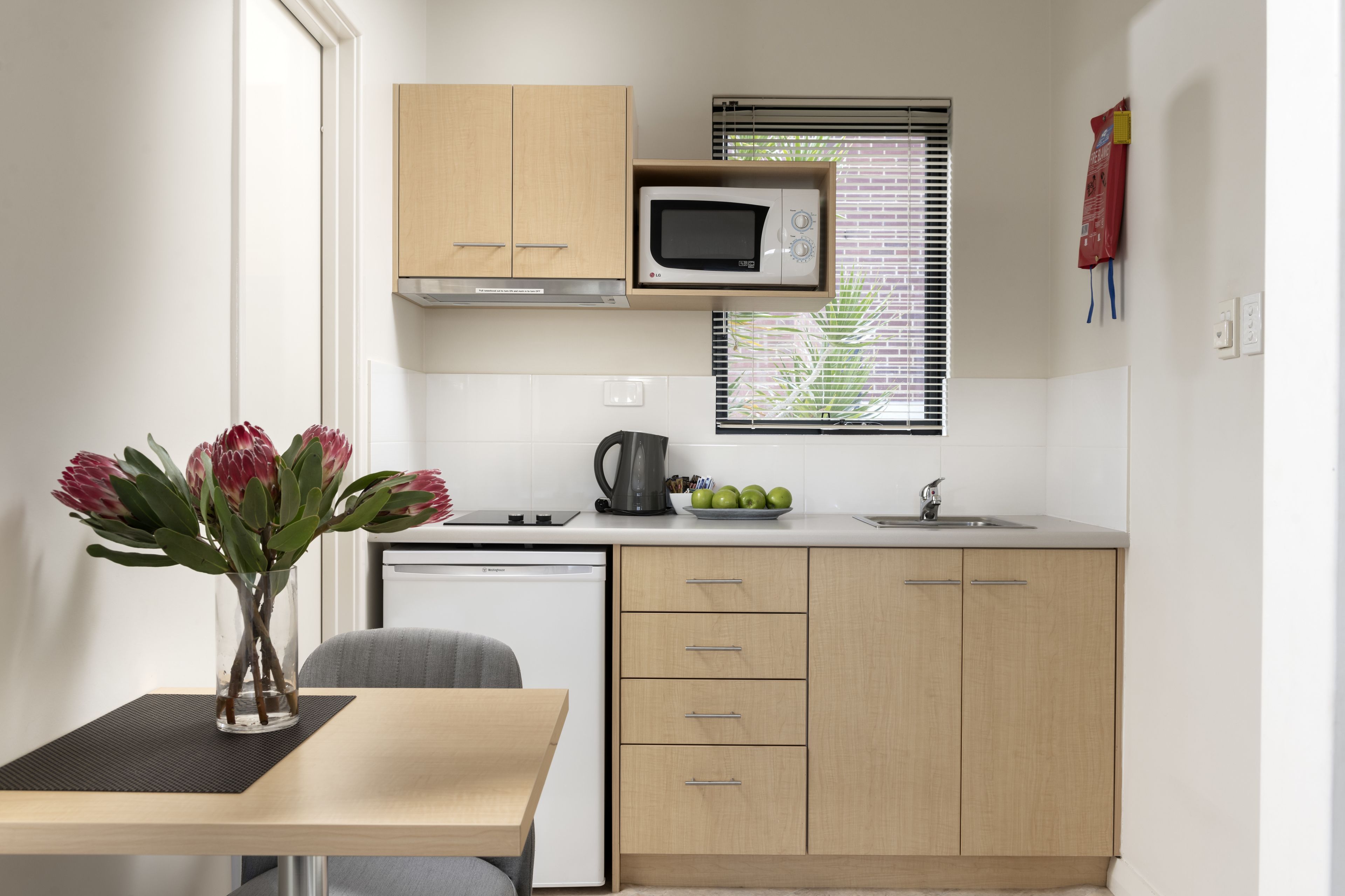 Una cocina compacta con armarios de madera, un microondas, un lavamanos y un ventanal con persianas.