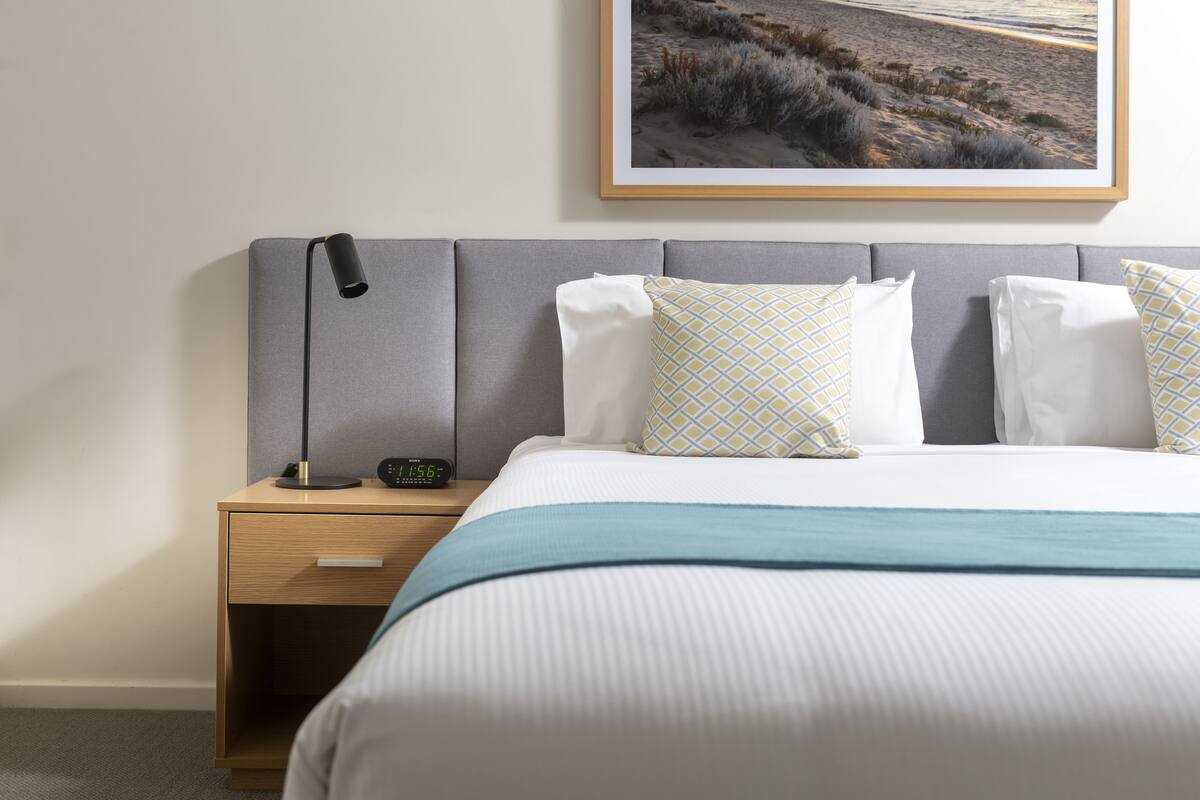 Un lit bien fait, recouvert d’une couvre-lit blanche et bleue, avec une tête de lit grise et des coussins à motifs. On aperçoit une table de chevet avec une lampe et une horloge.