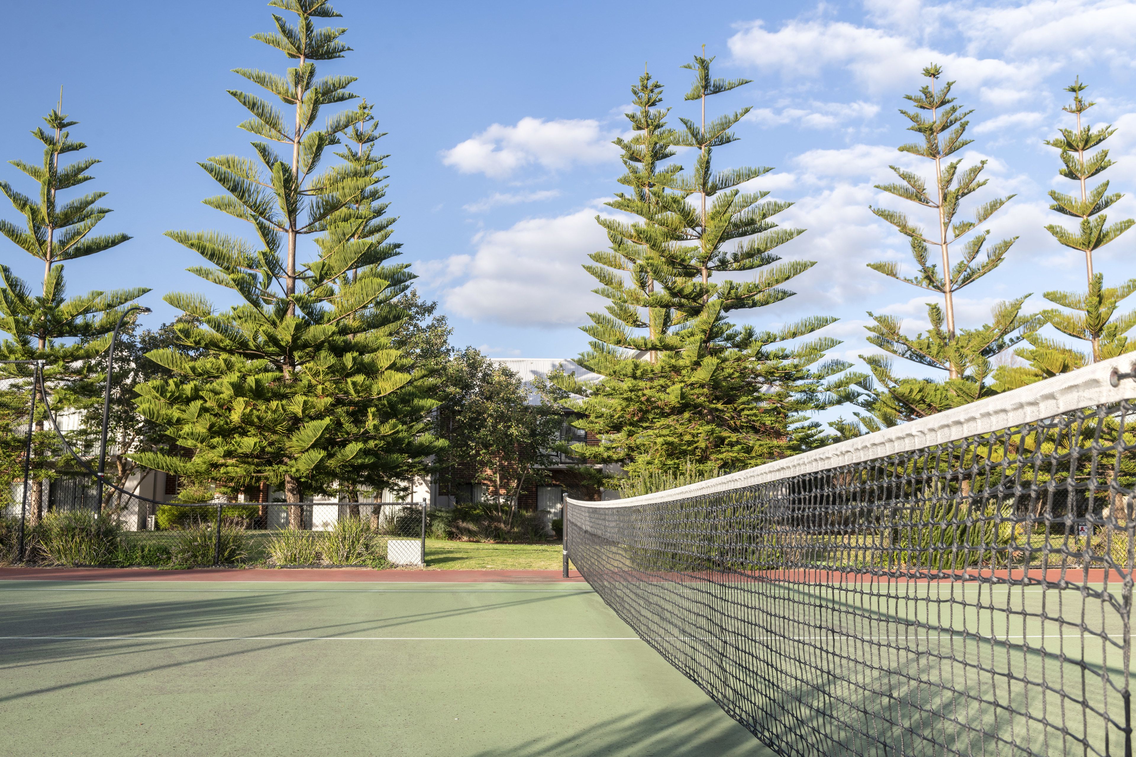Un court de tennis avec un filet, entouré de grands arbres et avec un bâtiment en arrière-plan.