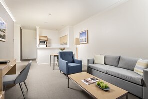A modern living room with a grey sofa, a blue armchair, a wooden coffee table with a magazine and apples, a desk with a laptop, and a kitchen area in the background.