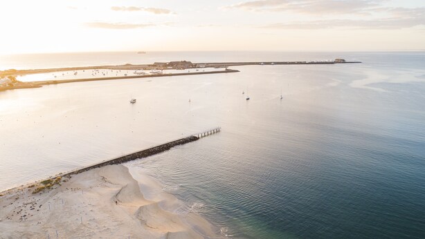 Vue aérienne d’une zone côtière avec une plage de sable, une eau calme et une digue qui s’avance dans la mer.