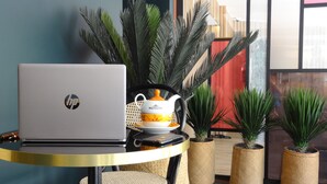 Un ordenador portátil sobre una mesa redonda, una tetera y plantas en macetas en una habitación moderna con vistas al exterior.