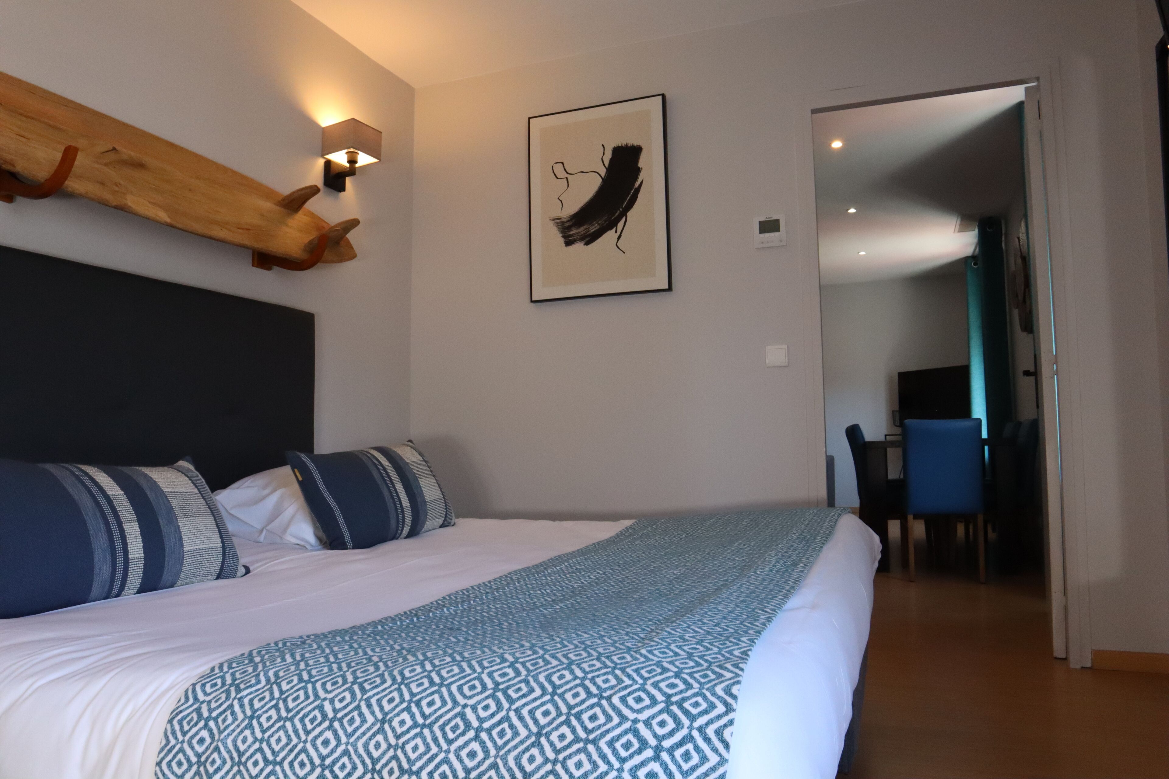 A hotel room with a large bed, a wooden headboard, a wall-mounted light, a framed abstract artwork, and a desk with a chair in the background.