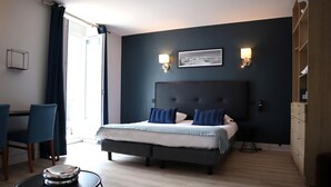 Un dormitorio moderno con un cabecero oscuro, una cama con ropa de cama azul y blanca, dos lámparas fijadas a la pared y una mesita de noche con un libro.