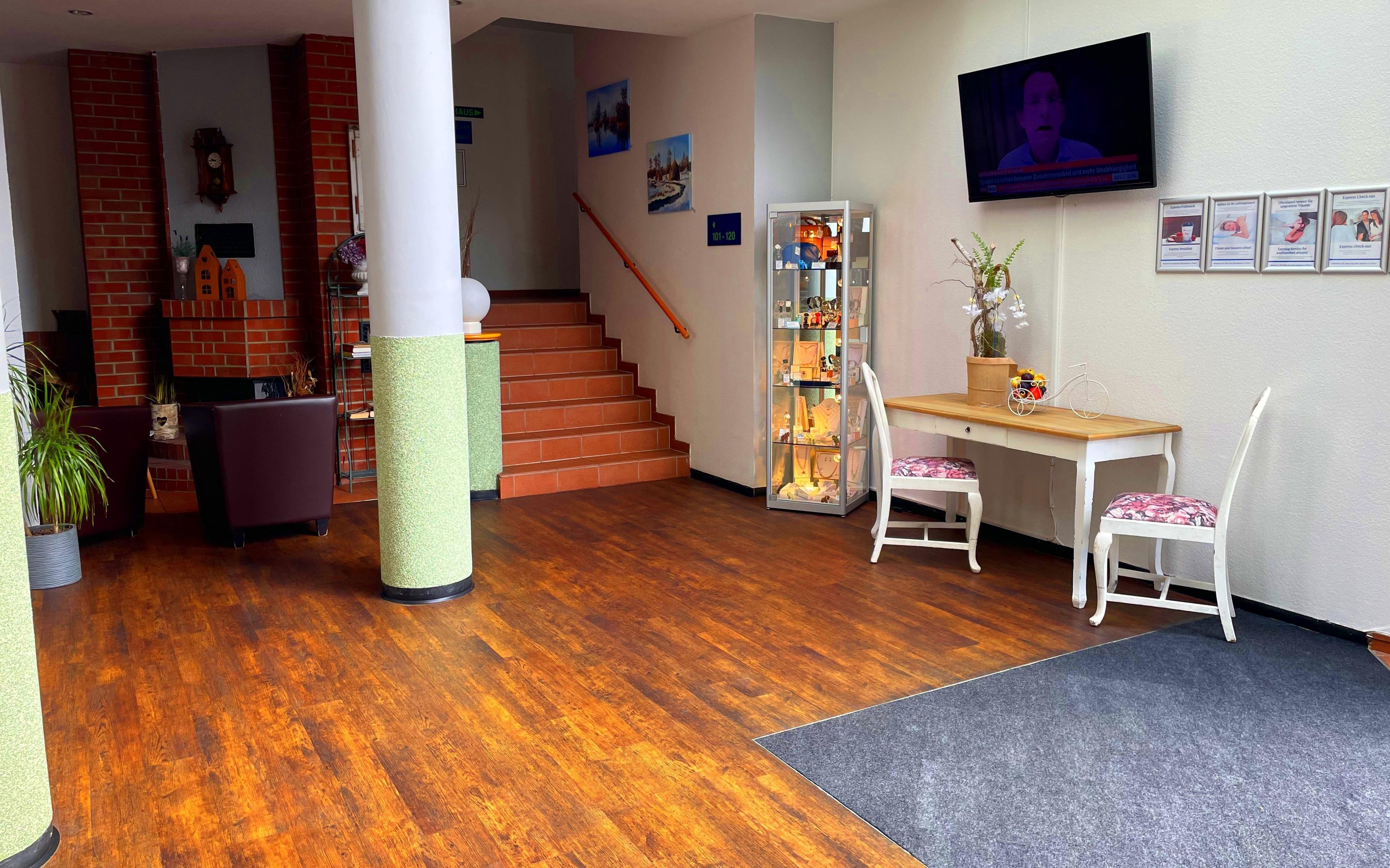 A hotel lobby with a wooden floor, a reception desk, a television mounted on the wall, and a staircase leading to another floor.