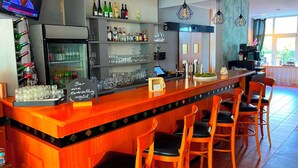 Zona de bar con mostrador de madera, taburetes y estanterías llenas de botellas y vasos.