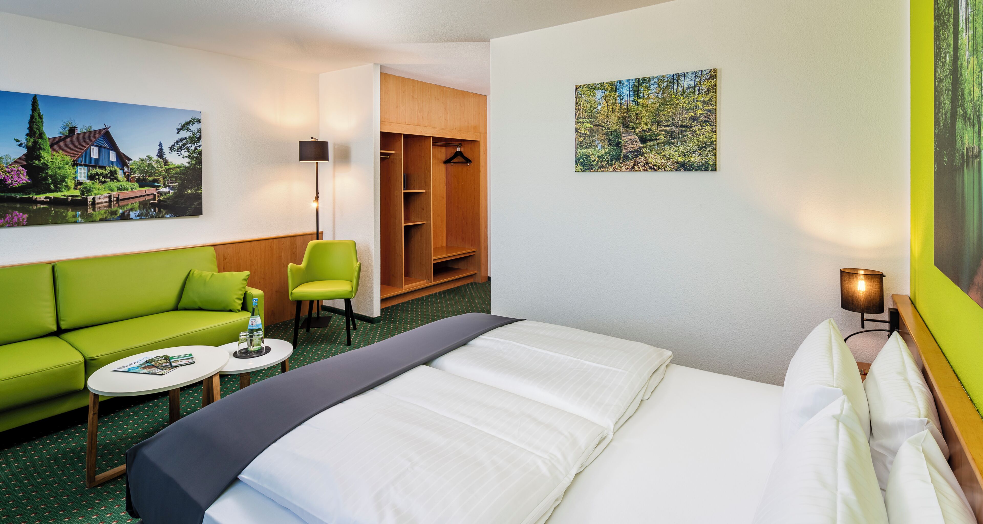 A hotel room with a bed, a green sofa, a small table, a chair, a wardrobe, and a painting on the wall.