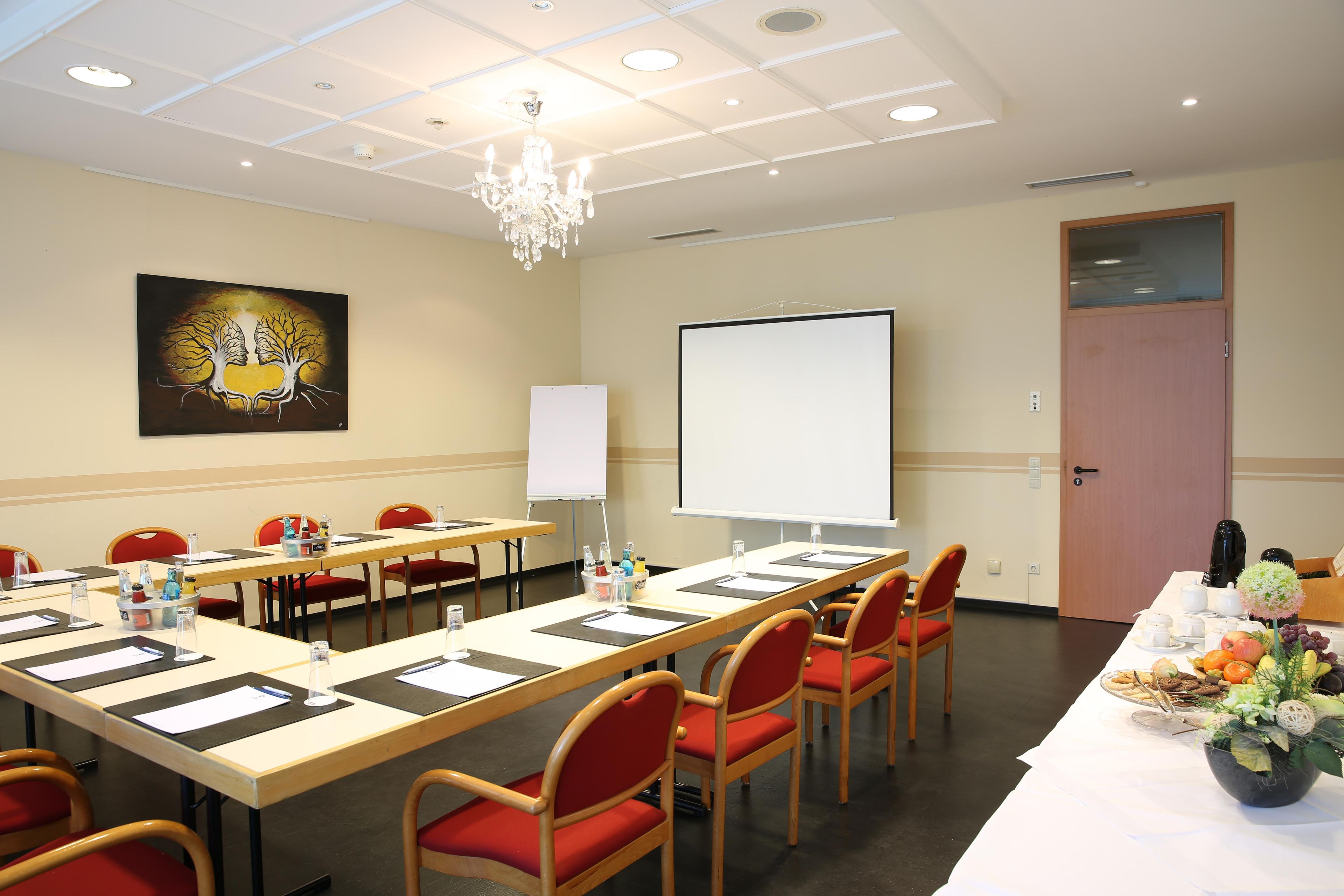 A conference room with a long table, red chairs, a projector screen, and a painting on the wall.