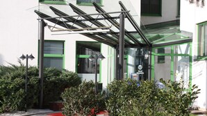 Entrada de hotel con toldo de cristal, ventanas con marco verde y alfombra roja que conduce a la puerta.