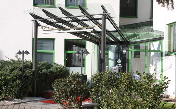 Entrada de hotel con toldo de cristal, ventanas con marco verde y alfombra roja que conduce a la puerta.