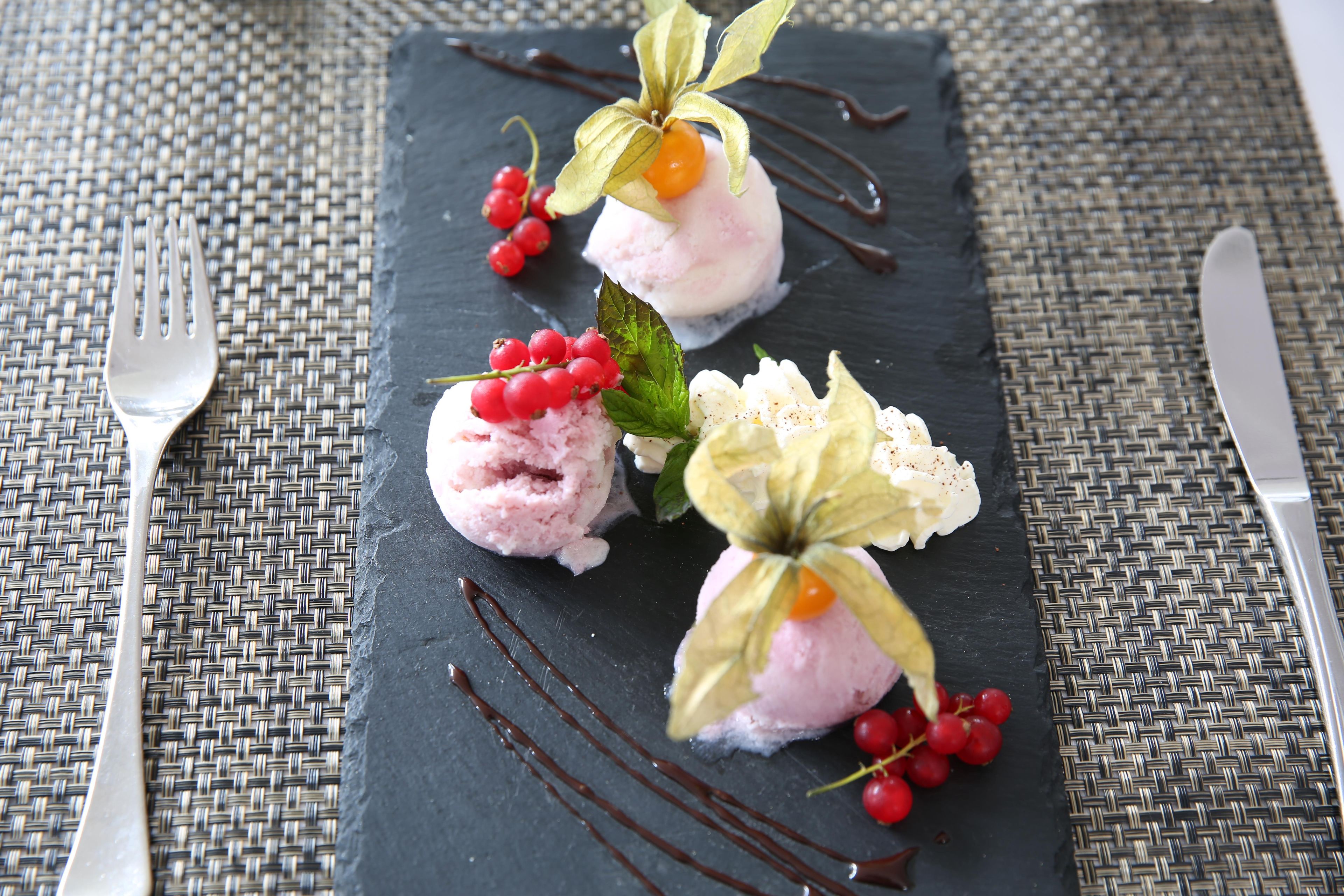 Un plato de postre con tres bolas de helado rosa, adornado con bayas rojas y hojas verdes, sobre un plato de pizarra oscura.