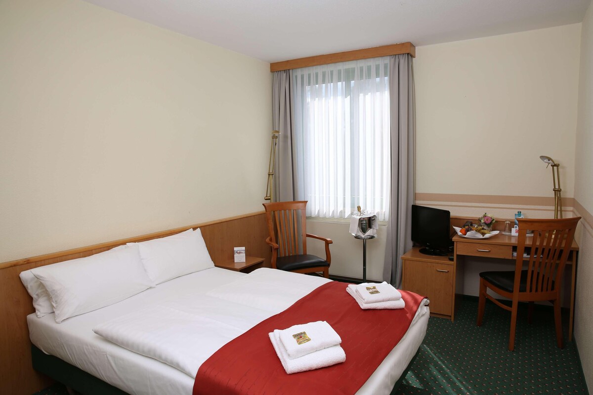 Habitación de hotel con cama, escritorio, silla, televisor y ventana con cortinas.