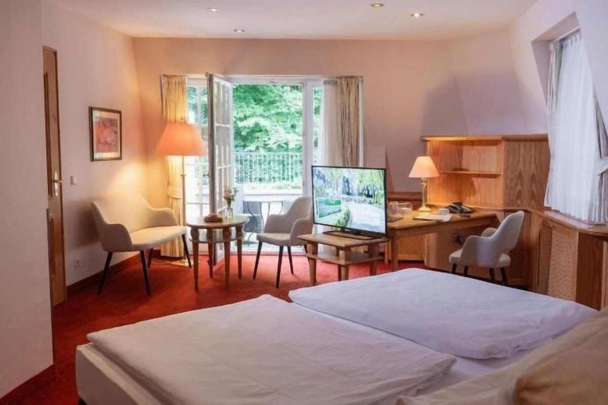 A hotel room with a bed, a desk, a television, and a view of greenery outside.