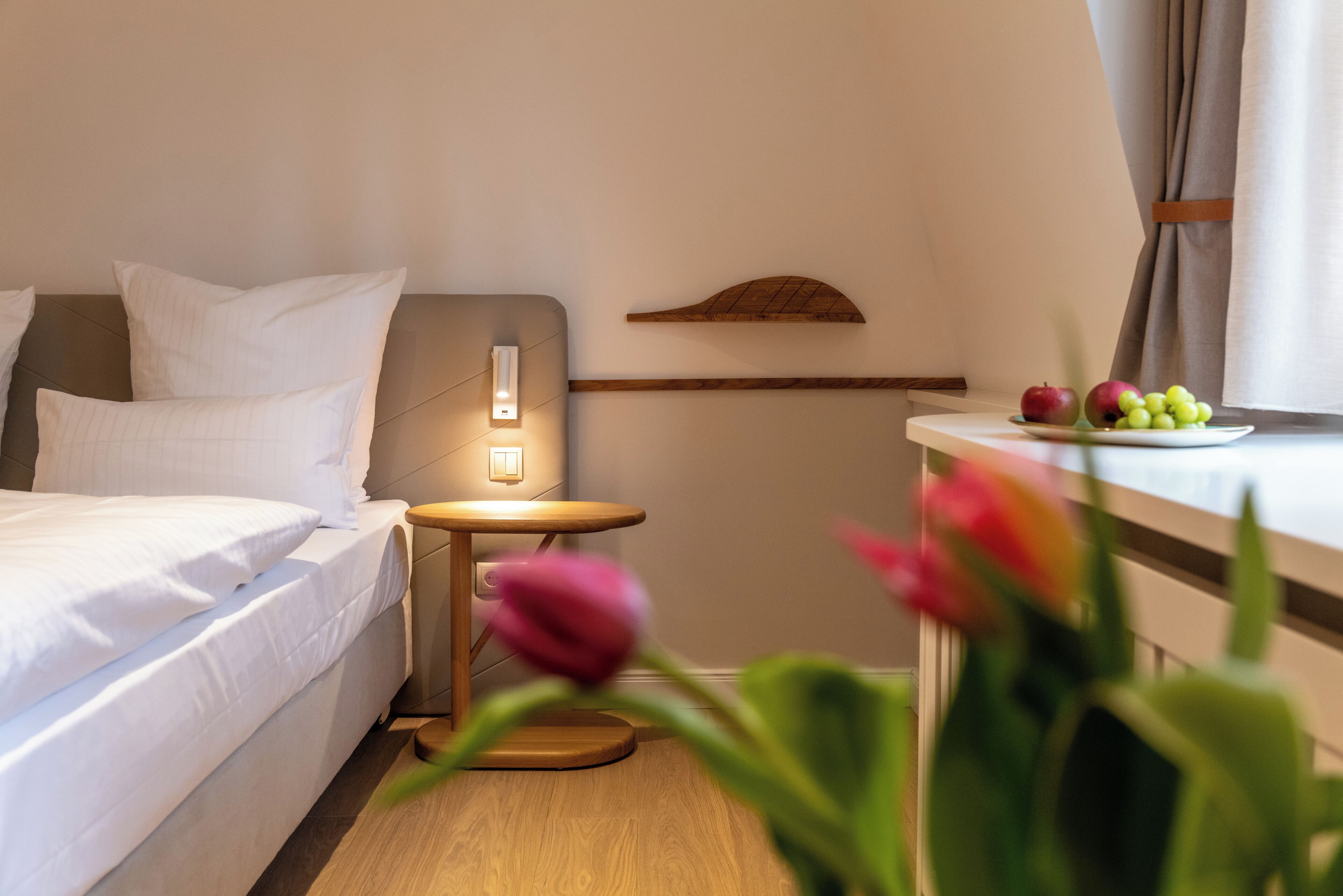 Habitación de hotel con cama, mesita de noche, luz de pared y una pequeña mesa con fruta y flores.