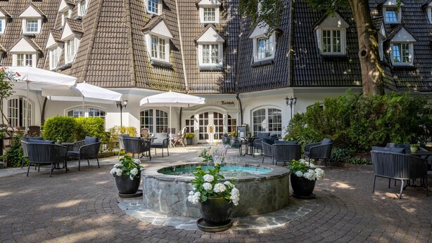 A courtyard with outdoor seating, a fountain, and a building with a tiled roof and arched windows. - Waldhaus Reinbek (Reinbek)
