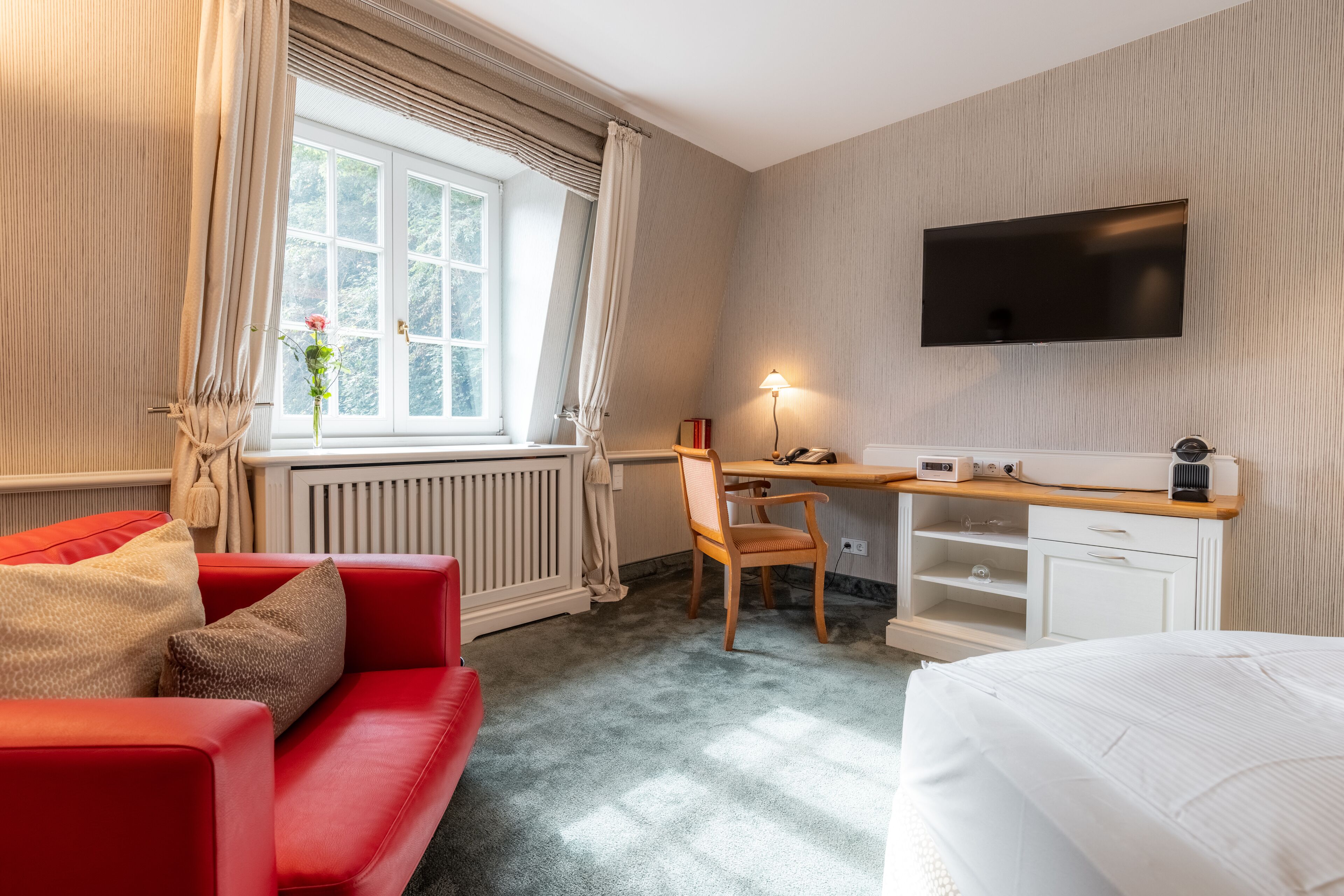 A hotel room with a red sofa, a white bed, a desk with a computer and lamp, a TV on the wall, and a window with curtains.