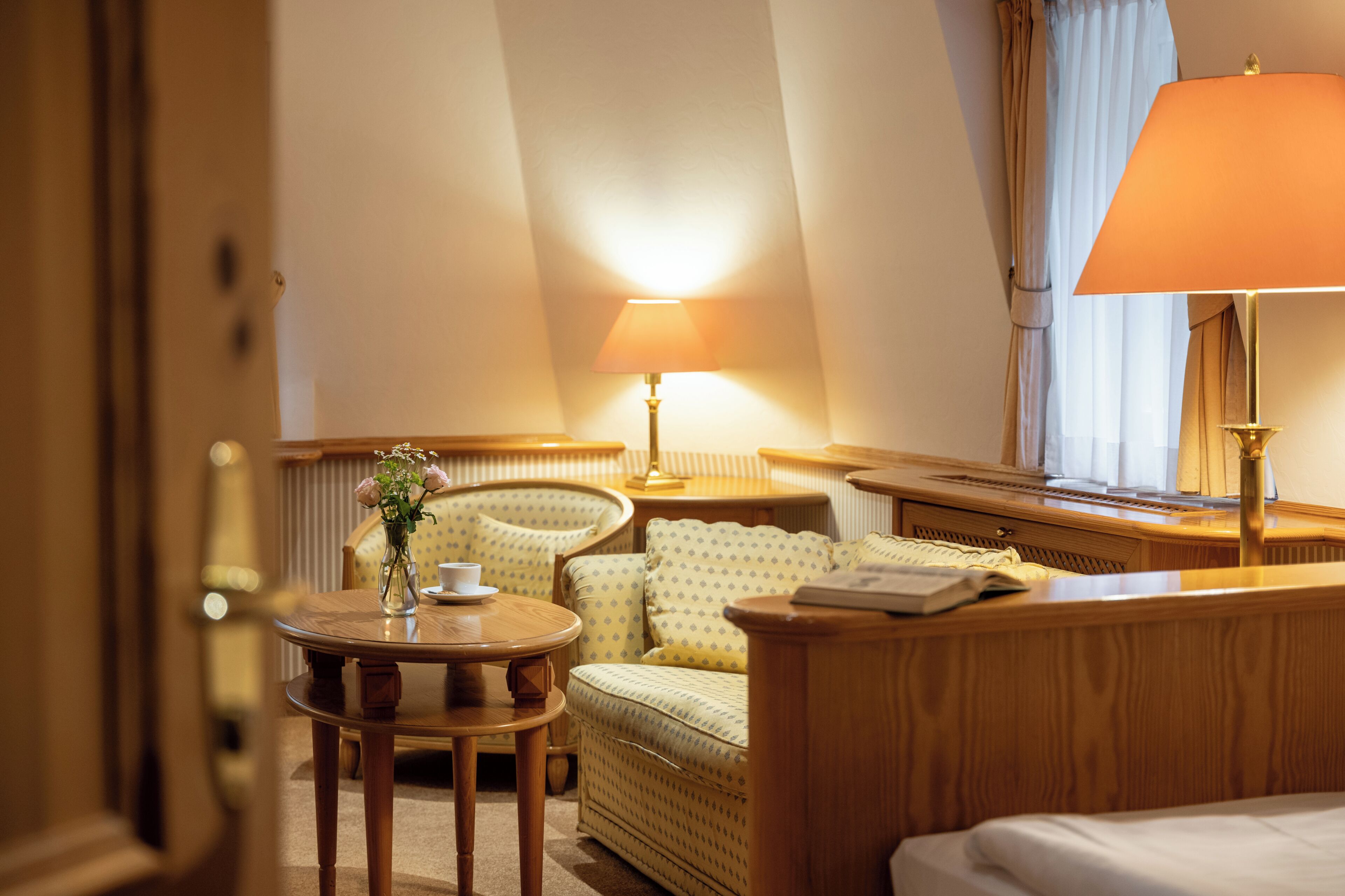 A hotel room with a bed, a round table with a vase of flowers, a sofa, a lamp, and a window with curtains.