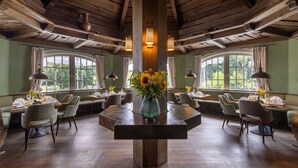 A dining area with a central round table surrounded by chairs, wooden beams on the ceiling, and large windows with curtains. - Waldhaus Reinbek (Reinbek)