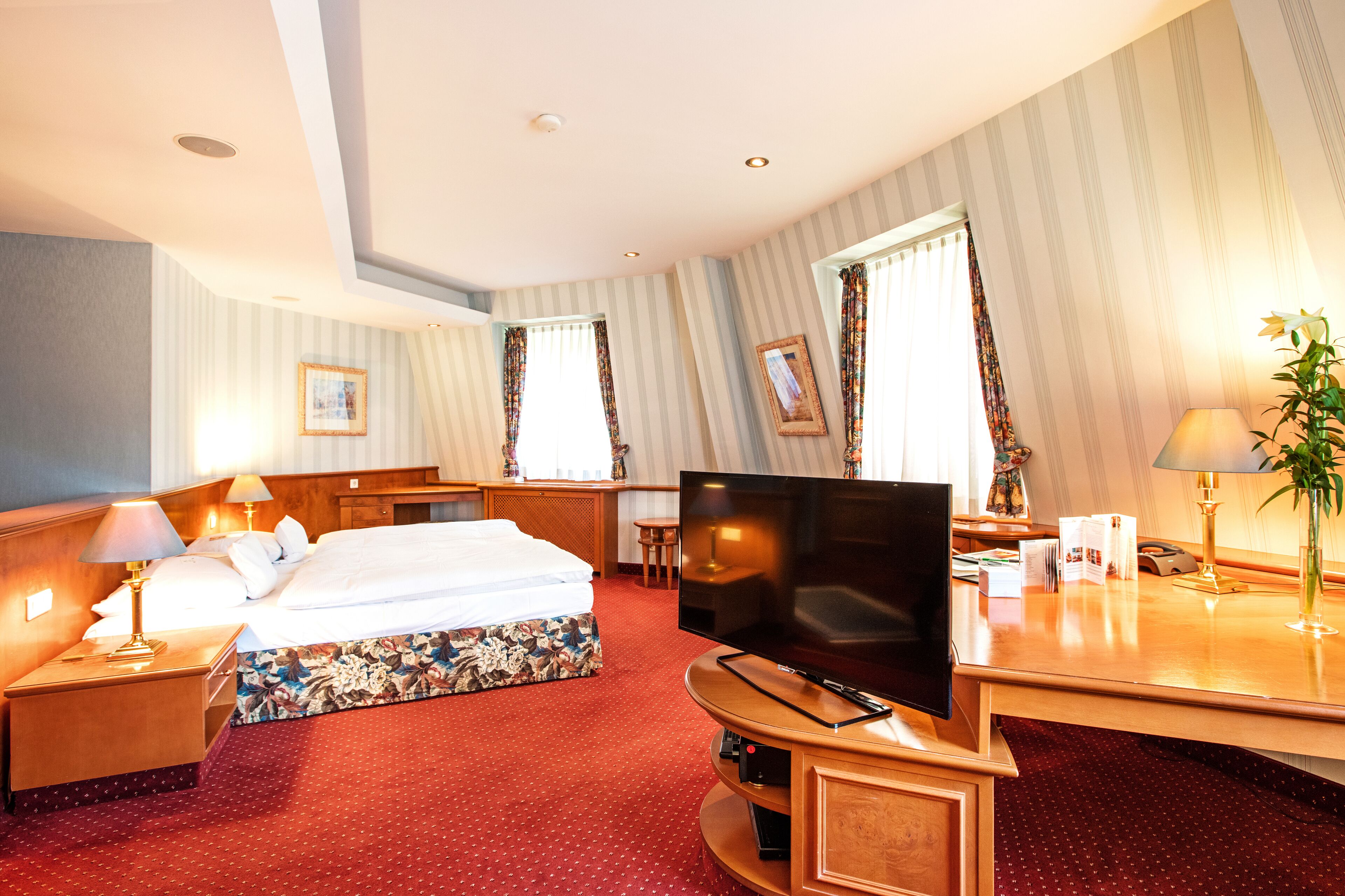 Habitación de hotel con una cama, un escritorio con televisor, una lámpara y una planta.