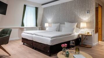 Quarto de hotel com cama de casal, duas mesas de cabeceira com abajures, piso de madeira, uma cadeira verde e uma pequena mesa com um vaso e copos.