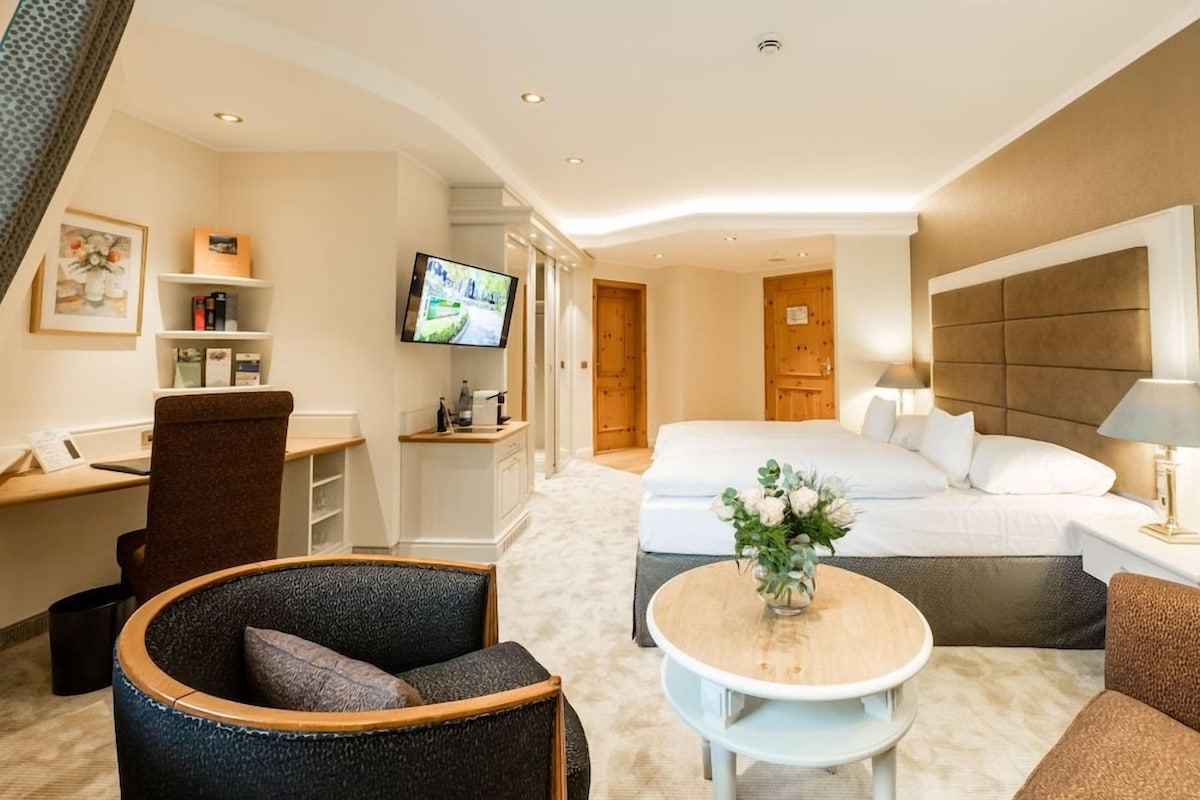 A hotel room with a bed, a desk, a chair, a TV, and a small table with a vase.